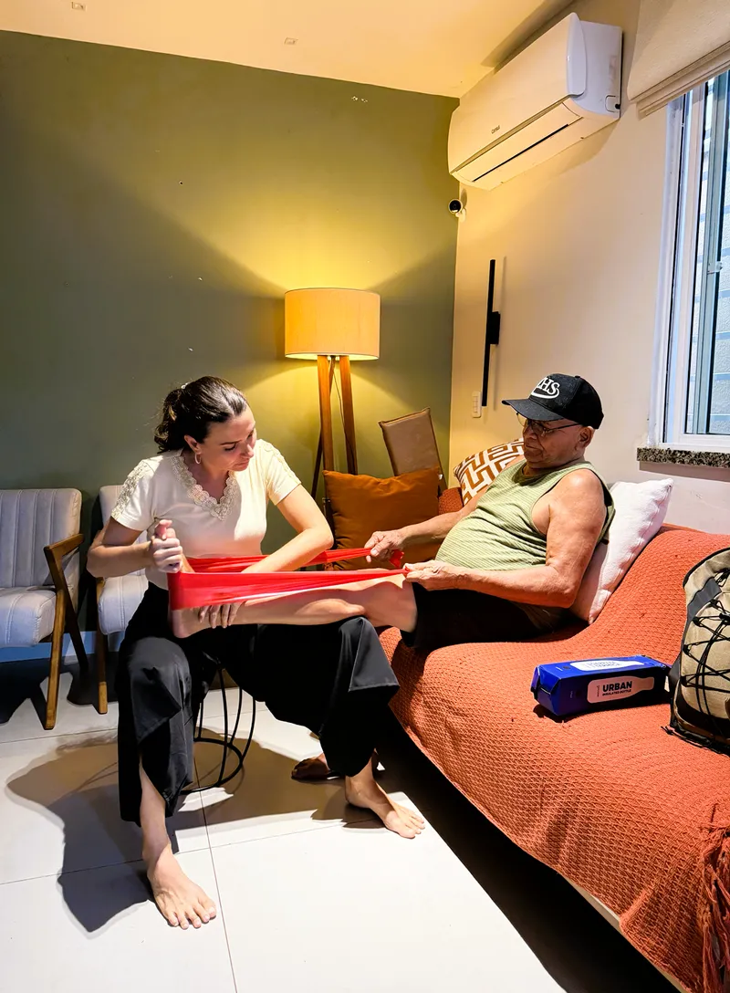 Fisioterapeuta conduzindo exercício com faixa elástica na perna de um senhor sentado no sofá.